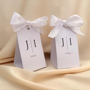 May include: Two white favour boxes with white satin bows. Each box has the initials "J I" and the date "01.08.26" printed in black. The boxes are on a cream-coloured satin surface.