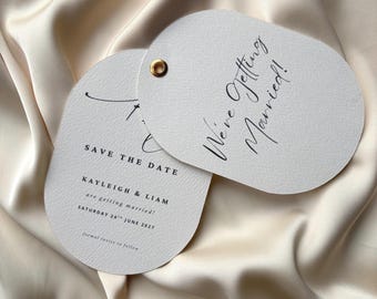 May include: Two off-white, oval-shaped wedding announcement cards with gold eyelets. One card reads "We're Getting Married!" in elegant script. The other card says "Save the Date" with names and a wedding date.
