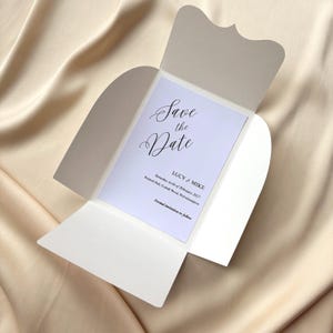 Pode incluir: Um convite de casamento branco com uma aba decorativa, com o texto "Save the Date" em escrita elegante. O convite inclui os nomes "LUCY & MIKE" e detalhes do evento. O fundo é um tecido macio de cor creme.