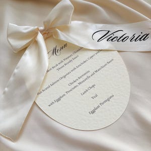 Może przedstawiać: Kremowa karta menu z satynową wstążką zawiązaną w kokardę. Menu jest okrągłe i zawiera listę dań. Na wstążce wydrukowano imię "Victoria". Menu znajduje się na kremowej satynowej powierzchni.