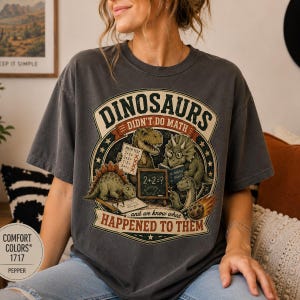 May include: A charcoal gray t-shirt featuring a graphic of three dinosaurs with a chalkboard and books. The text reads: DINOSAURS DIDN'T DO MATH and ...and we know what HAPPENED TO THEM. The design includes a T-Rex, a Triceratops, and a Brachiosaurus.