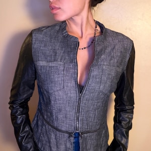 Può includere: Giacca grigia con maniche in pelle nera. La giacca ha una chiusura con cerniera e due piccole tasche sul petto. Le maniche sono in pelle nera liscia. Un capo di abbigliamento esterno elegante.