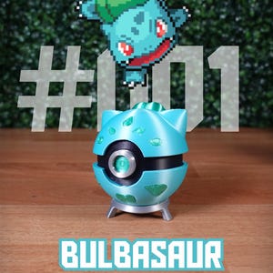 Puede incluir: Un objeto decorativo turquesa con el tema de Bulbasaur, con una banda negra y detalles plateados. El diseño incluye una imagen pixelada de Bulbasaur y el texto "#01 BULBASAUR". El objeto está sobre una superficie de madera.