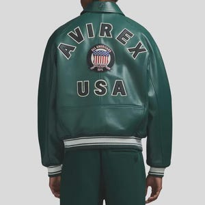 May include: Green leather jacket with white and black striped cuffs and hem. The back of the jacket features the word "AVIREX" in white, arched above a circular patch with the American flag and the words "ALL AMERICANS" and "USA".