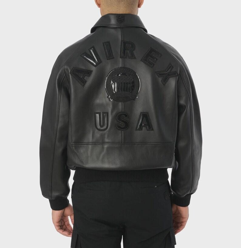 May include: Black leather bomber jacket with the word "AVIREX" arched across the back, above a circular emblem and the word "USA". The jacket has a ribbed hem and cuffs, and a collared neckline.
