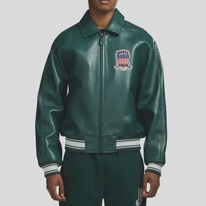 May include: A green leather jacket with a zip-up front, a collar, and a patch on the chest. The jacket has white and gray striped cuffs and a matching hem. The patch features a shield design with the text "AVIREX".