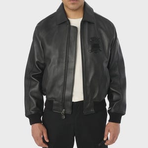 May include: Black leather jacket with a collared neckline, a zipper closure, and a black emblem on the chest. The jacket has ribbed cuffs and a ribbed hem. The model is wearing a white shirt underneath.