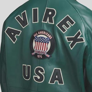 May include: A green leather jacket with white and black lettering and patches. The back of the jacket features the word "AVIREX" and "USA" in large white letters with black outlines. A circular patch displays the American flag and the words "ALL AMERICANS ESTABLISHED 1975".