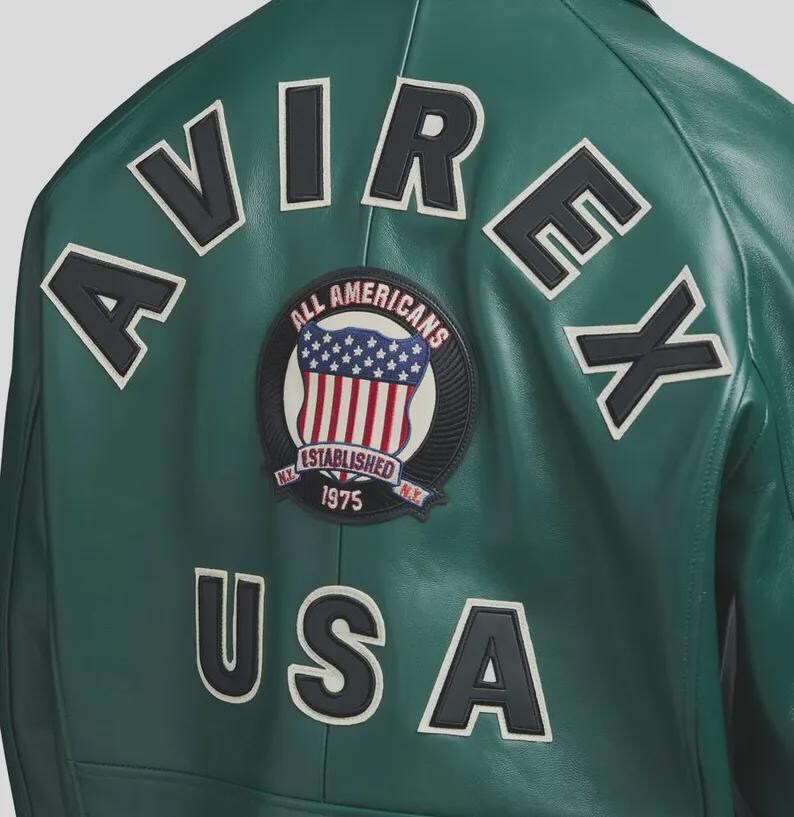 May include: A green leather jacket with white and black lettering and patches. The back of the jacket features the word "AVIREX" and "USA" in large white letters with black outlines. A circular patch displays the American flag and the words "ALL AMERICANS ESTABLISHED 1975".