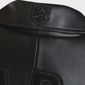 May include: Close-up of a black leather jacket. The jacket features a small, embroidered emblem on the collar and large, stitched letters on the lower portion. The leather has a smooth, matte finish. The jacket appears to be new.