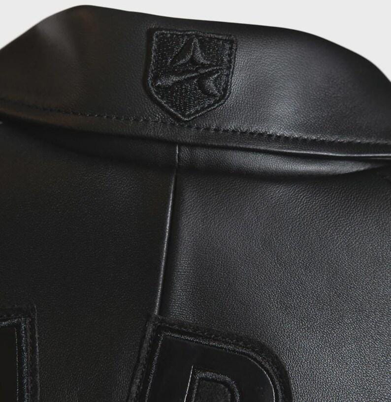 May include: Close-up of a black leather jacket. The jacket features a small, embroidered emblem on the collar and large, stitched letters on the lower portion. The leather has a smooth, matte finish. The jacket appears to be new.