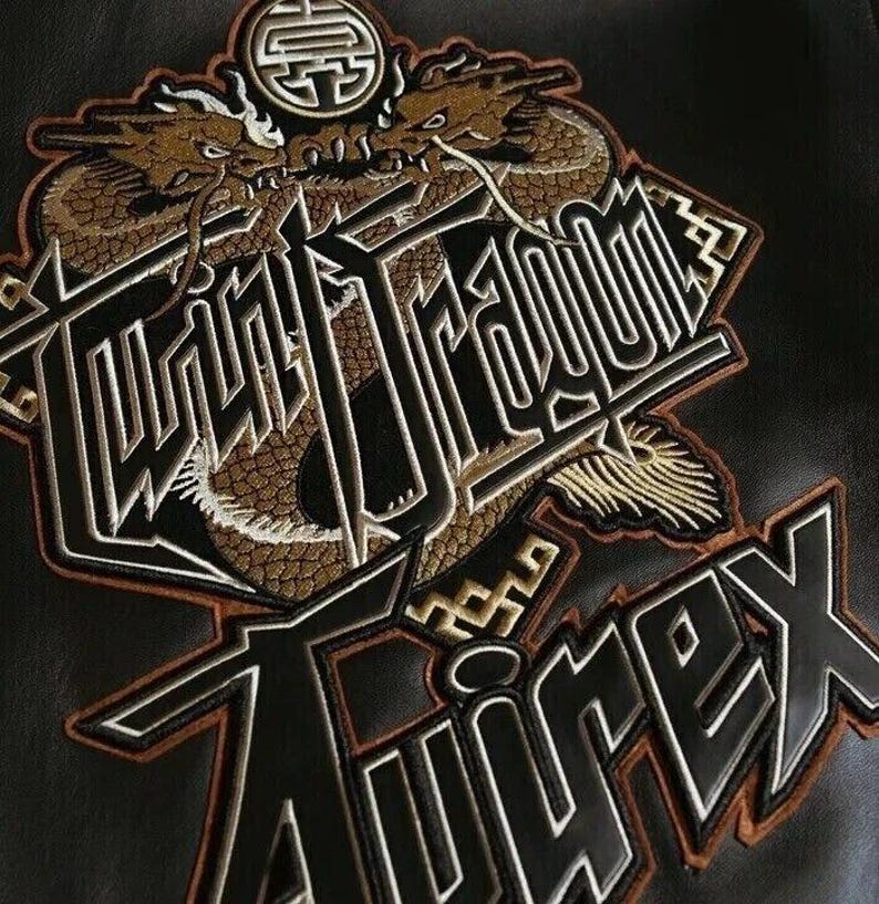 May include: Embroidered patch featuring two golden dragons above the words "Twin Dragon" and "Avirex" in a bold, stylized font. The patch has a black background with brown and white accents, creating a striking contrast.