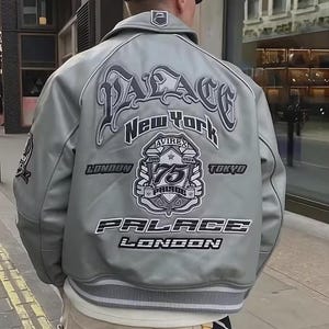 Pode incluir: Um casaco estilo universit&aacute;rio cinzento-claro com um grande texto bordado nas costas que diz Palace, New York, London e Tokyo. O design inclui um emblema central com o n&uacute;mero 75 e a marca Avirex, com acabamentos em malha canelada &agrave;s riscas.