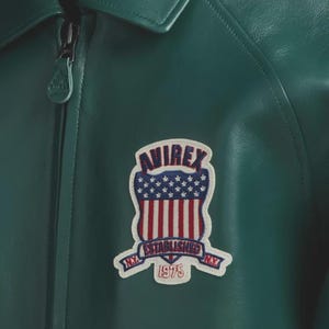 May include: Close-up of a green leather jacket with a zipper and a patch. The patch features the word "AVIREX" above a design of the American flag and the words "ESTABLISHED 1975 N.Y."