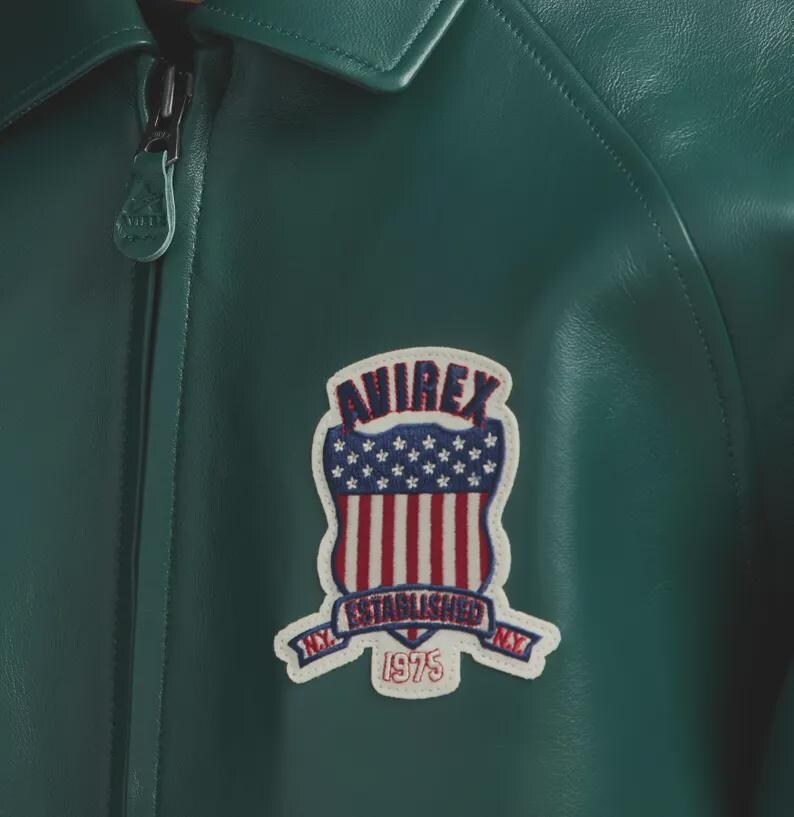 May include: Close-up of a green leather jacket with a zipper and a patch. The patch features the word "AVIREX" above a design of the American flag and the words "ESTABLISHED 1975 N.Y."