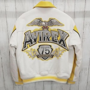 May include: A white leather jacket with yellow accents and a yellow collar. The back features a large embroidered graphic of an eagle above the text AVIREX and a shield with the number 75 and the text AVIATOR'S KING in black and yellow.