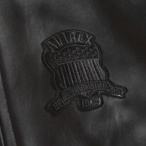 May include: Close-up of a black leather jacket featuring a textured patch. The patch is a shield shape with the text "AVIREX ESTABLISHED NYT 1975" in a raised, tonal design. The jacket's leather has a subtle grain.
