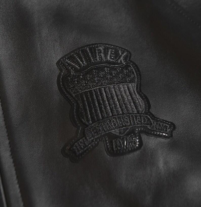 May include: Close-up of a black leather jacket featuring a textured patch. The patch is a shield shape with the text "AVIREX ESTABLISHED NYT 1975" in a raised, tonal design. The jacket's leather has a subtle grain.