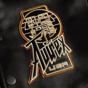 May include: Black and gold embroidered patch with the word "Timex" and "USA" on a black leather jacket. The patch has a circular design with text and a rectangular base.