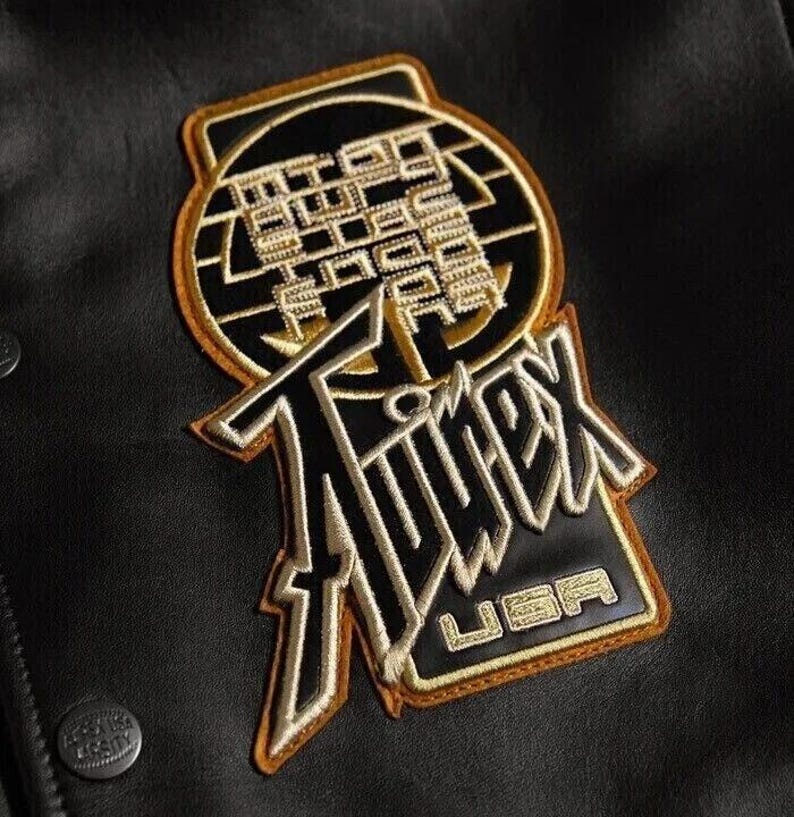 May include: Black and gold embroidered patch with the word "Timex" and "USA" on a black leather jacket. The patch has a circular design with text and a rectangular base.