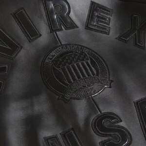 May include: Close-up of a black leather jacket with embossed lettering and a circular emblem. The emblem reads "All American" and "Established 1975". The jacket has a classic, vintage aesthetic.