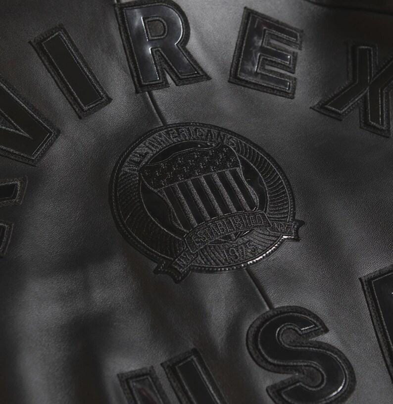 May include: Close-up of a black leather jacket with embossed lettering and a circular emblem. The emblem reads "All American" and "Established 1975". The jacket has a classic, vintage aesthetic.