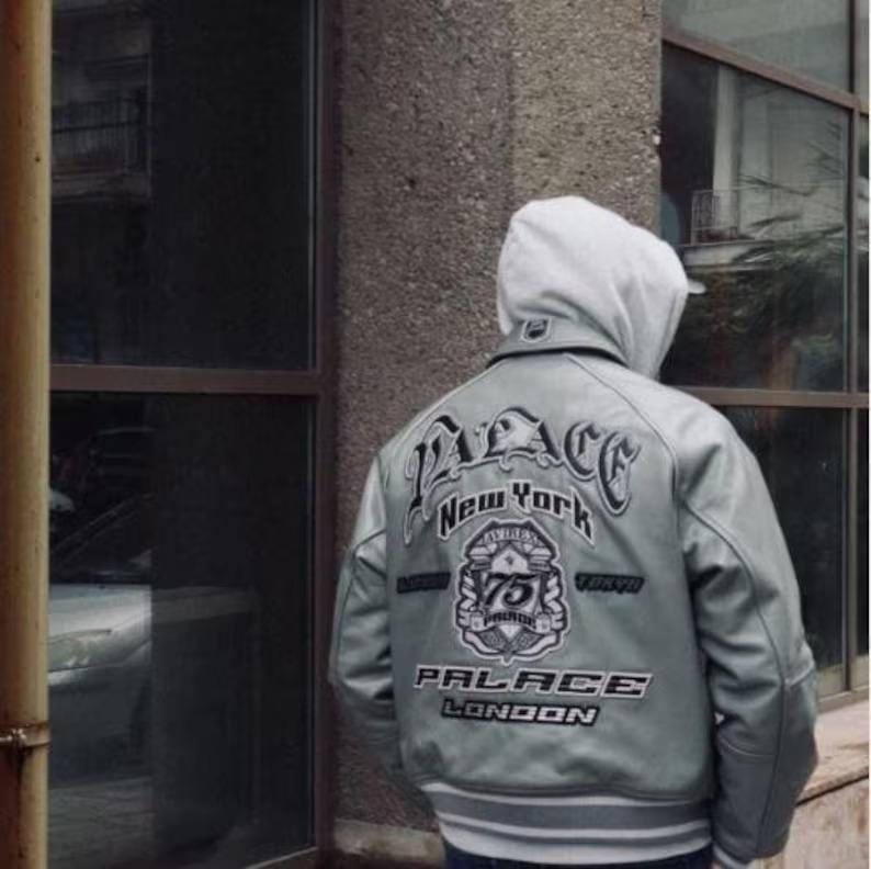 Pode incluir: Uma jaqueta universit&aacute;ria de couro cinza-claro com um moletom branco com capuz por baixo. As costas da jaqueta exibem texto bordado Palace New York, Tokyo e London, com um emblema central apresentando o n&uacute;mero 75.