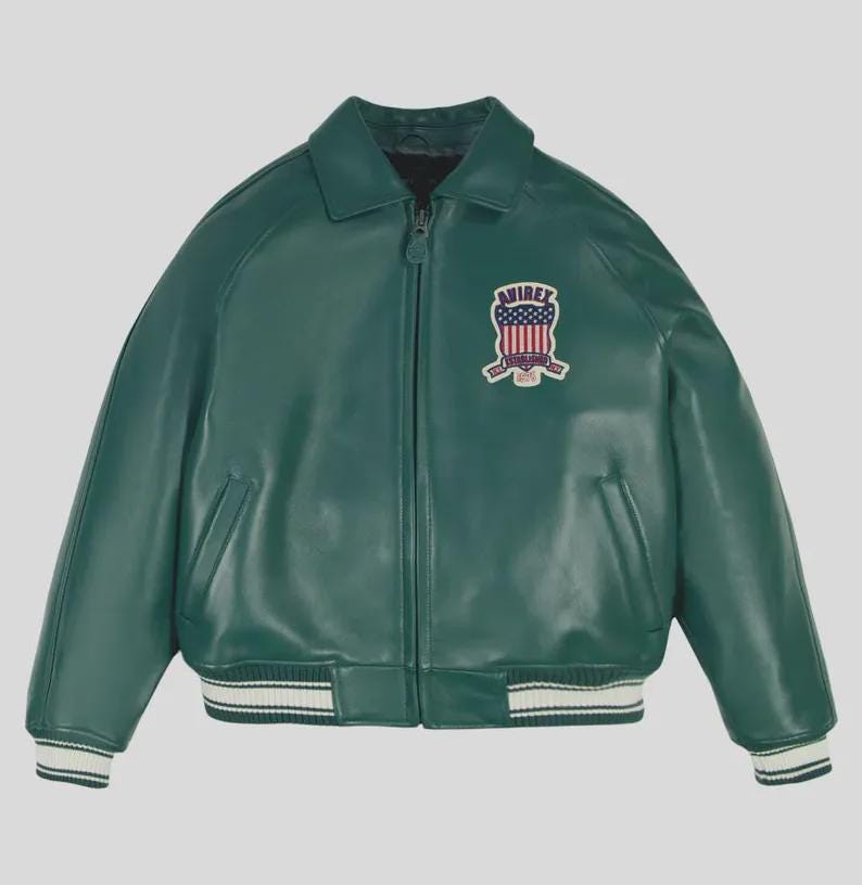 May include: A green leather jacket with a zip-up front, collar, and a patch on the upper left chest. The patch features a shield design with red, white, and blue elements. The jacket has white and black striped cuffs and hem.