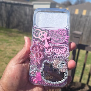 Könnte beinhalten: Transparente Handyhülle mit rosa und silbernen Strasssteinen. Sie zeigt ein Hello Kitty-Motiv, das Wort "Chanel" und andere dekorative Elemente. Die Hülle wird vor einem sonnigen Hintergrund im Freien gehalten.