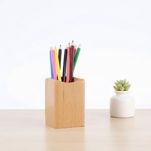 May include: A wooden pencil holder filled with colorful pencils. The pencils are various colors, including blue, pink, yellow, black, and green. A small white vase with a green succulent plant sits to the right of the pencil holder.