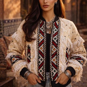 May include: A cream-coloured bomber jacket with detailed geometric patterns in red, green, and yellow. The jacket has fringe detailing and sequin embellishments. Black cuffs and collar provide contrast. The text "ATLAS TREASURE" is visible on the top left.