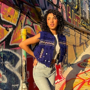 Könnte beinhalten: Eine Frau trägt eine blaue Weste mit Fransen und gestickten Details, ein weißes Oberteil und eine hellblaue Jeans. Der Hintergrund zeigt farbenfrohe Graffiti-Kunst an einer Wand.