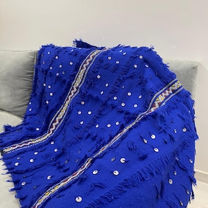 May include: A vibrant royal blue textile blanket featuring a textured surface with scattered silver sequins and tufted fringe details. The fabric is accented by two vertical woven bands displaying a geometric pattern in red, white, and yellow.