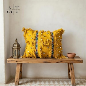 May include: A rectangular yellow decorative pillow with fringe and silver embellishments. The pillow is on a wooden bench with a small lantern and a terracotta bowl. The text "ATLAS TREASURE" is in the upper left corner.