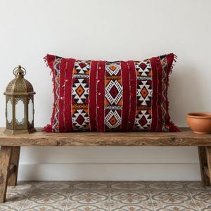 May include: A rectangular throw pillow featuring a deep red base with vertical stripes and intricate geometric patterns in white, black, and orange. The pillow is accented with small, reflective sequins and has frayed edges, displayed on a rustic wooden bench.