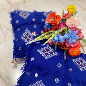Peut inclure: Textile bleu vif orné de motifs géométriques blancs et de franges. Un bouquet de fleurs colorées, dont des coquelicots orange, des œillets roses et des hortensias bleus, est posé sur le textile. Le motif présente des maisons stylisées.