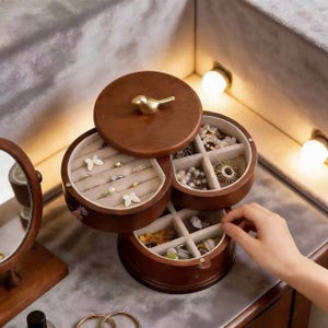 Può includere: Un portagioie in legno con tre scomparti rotanti. La scatola è marrone con una maniglia dorata a forma di uccello sul coperchio. I scomparti sono foderati in tessuto beige e contengono gioielli.