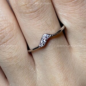 Puede incluir: Un delicado anillo de plata con un diseño ondulado, engastado con pequeñas piedras claras y brillantes. El anillo se muestra en un dedo, destacando su estilo elegante y minimalista.