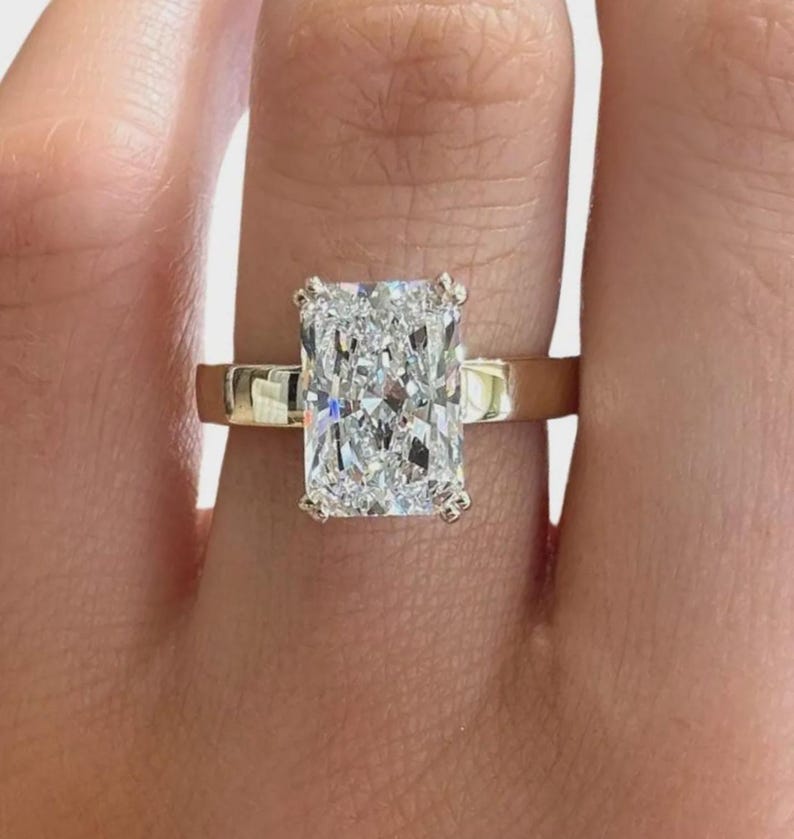 May include: A radiant-cut diamond ring with a gold band. The diamond is rectangular with a brilliant, multifaceted surface. The gold band is smooth and polished, creating a classic and elegant look. The ring is a symbol of luxury and commitment.