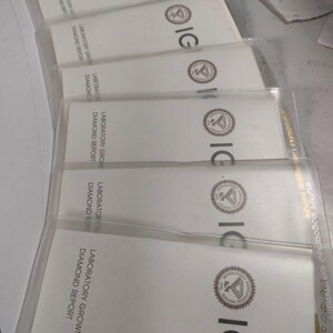 May include: A stack of white Laboratory Grown Diamond Report cards in clear plastic sleeves. The cards have the IGI logo and text. The cards are arranged in a slightly angled, overlapping pattern.