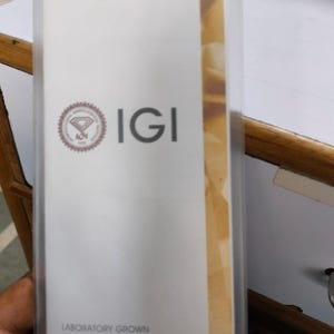 May include: A white IGI Laboratory Grown Diamond Report in a clear plastic sleeve. The report features the IGI logo and text. The report is on a white background with a tan stripe on the right side.