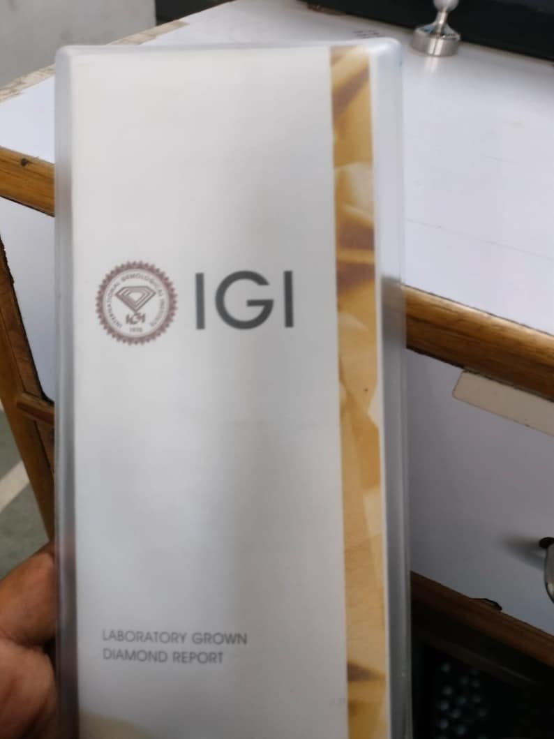 May include: A white IGI Laboratory Grown Diamond Report in a clear plastic sleeve. The report features the IGI logo and text. The report is on a white background with a tan stripe on the right side.