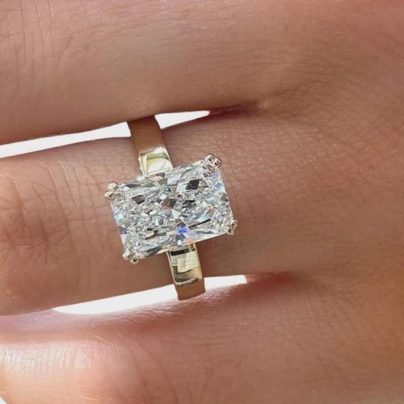 May include: A radiant-cut diamond ring with a gold band. The large, rectangular diamond sparkles with numerous facets, reflecting light. The ring is set on a gold band, showcasing a classic and elegant design.