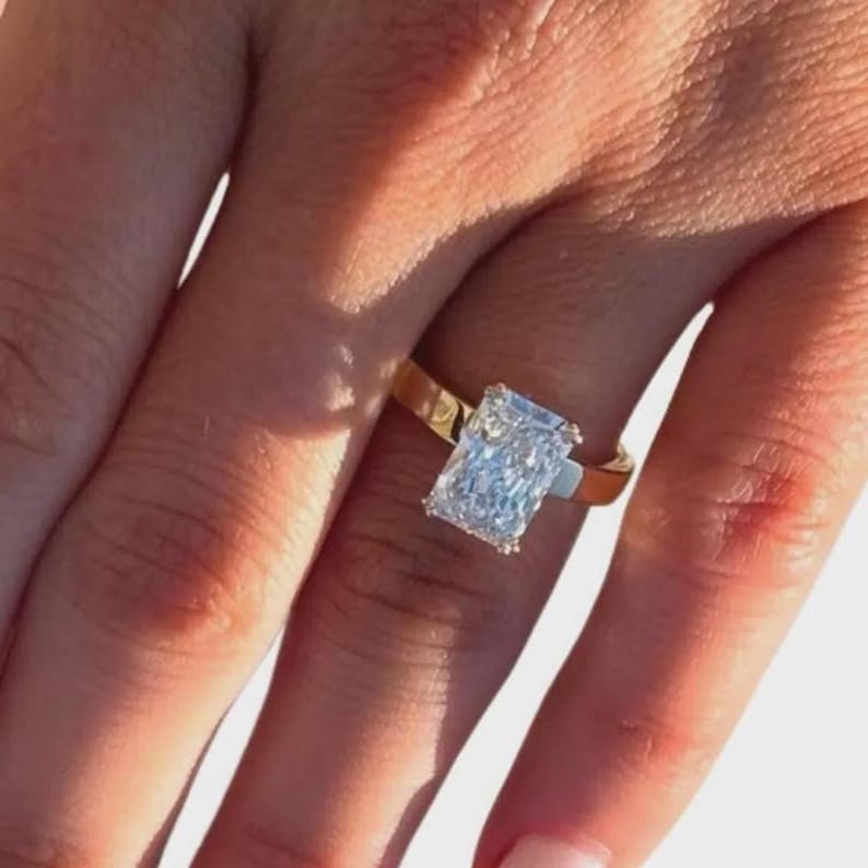 May include: A gold ring with a large, rectangular, clear gemstone. The ring is set on a finger, showcasing the design and sparkle of the stone. The band is a simple, polished gold.