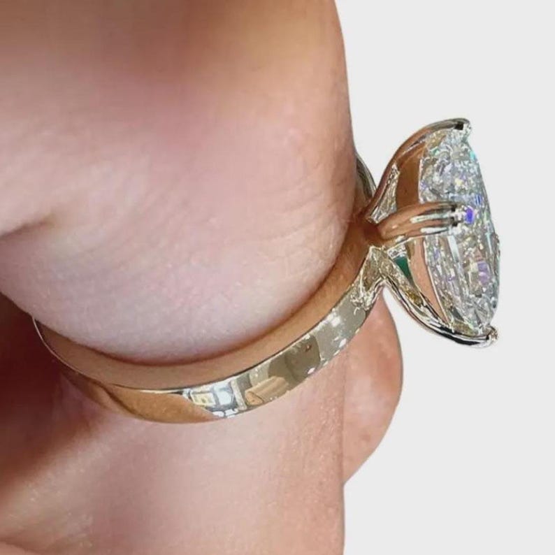 May include: A gold ring with a large, pear-shaped diamond. The diamond is clear and multifaceted, reflecting light. The ring's band is smooth and polished, with a classic design. The setting securely holds the diamond, showcasing its brilliance.