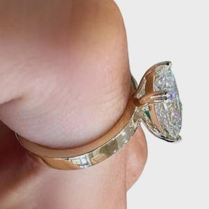 May include: A gold ring featuring a large, pear-shaped diamond. The diamond is clear and multifaceted, reflecting light. The band is smooth and polished, with a warm, golden hue. The ring is held by a person's fingers.