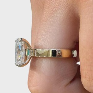 May include: A radiant-cut diamond ring with a gold band. The diamond is rectangular with rounded corners, set in a gold bezel. The ring is held by a person's fingers, showcasing the ring's design and sparkle.