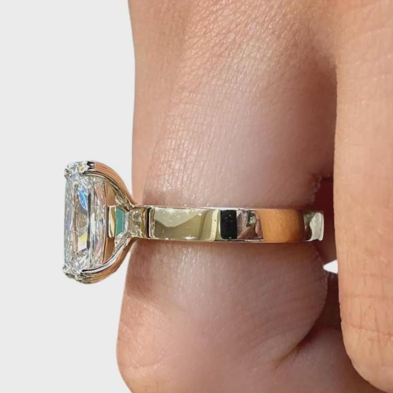 May include: A radiant-cut diamond ring with a gold band. The diamond is rectangular with rounded corners, set in a gold bezel. The ring is held by a person's fingers, showcasing the ring's design and sparkle.