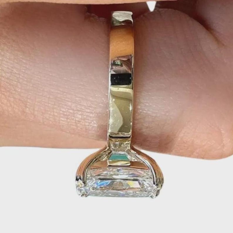 May include: A close-up of a gold ring with a large, rectangular, clear gemstone. The ring's band is a polished gold color, and the gemstone is set in a four-prong setting. The ring is shown against a neutral background.