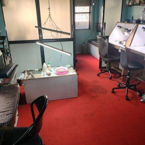 May include: An interior shot of a workshop with specialized equipment. The room features workbenches with built-in lighting, chairs, and a red carpet. A window provides natural light, and various tools and supplies are visible.